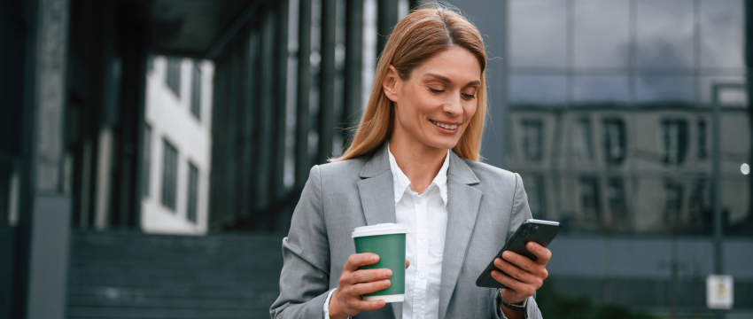 A professional woman in a business suit holds a coffee cup and gazes at her phone, illustrating a busy trading environment.